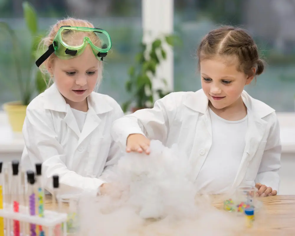 Two girls in white lab coats play with dry ice, fog spilling over the table.